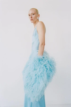 Model wearing a light blue slip dress adorned with fluffy feathers against a plain white studio wall.