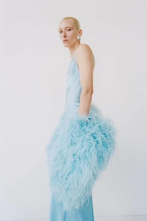 Model wearing a light blue slip dress adorned with fluffy feathers against a plain white studio wall.