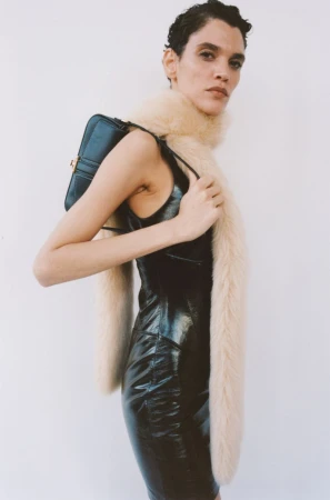 Model with short dark hair wearing a glossy black dress and furry cream scarf holding a black shoulder bag against white