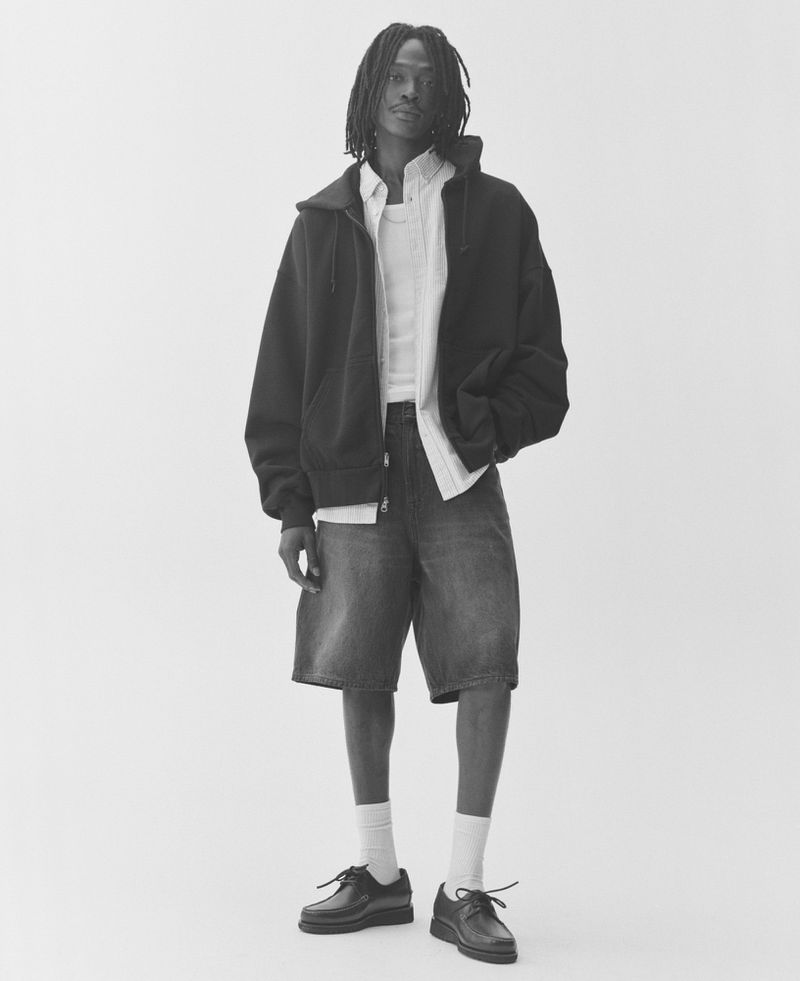 Black man wearing a dark zip-up hoodie over a white button-down shirt and denim shorts against a white backdrop.