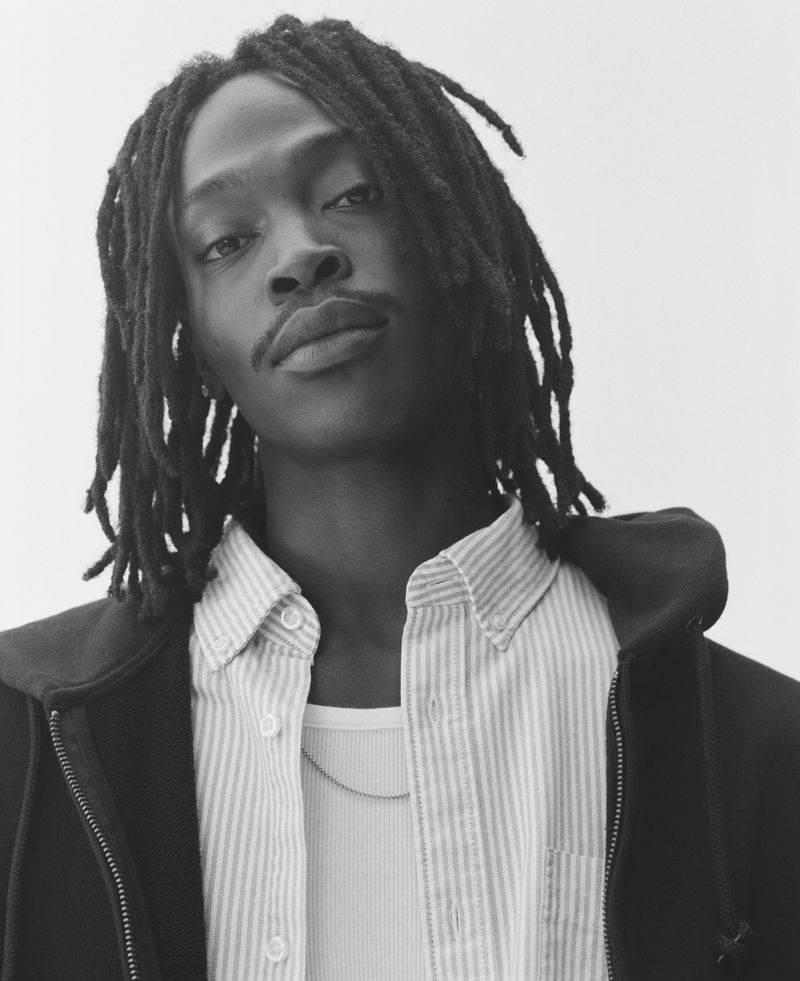 Portrait of a Black man with dreadlocks wearing a striped button-down shirt and a dark hooded jacket against a light background