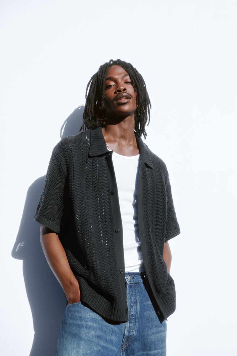 Model wearing a black knit polo shirt over a white tank top and blue jeans against a white seamless background.