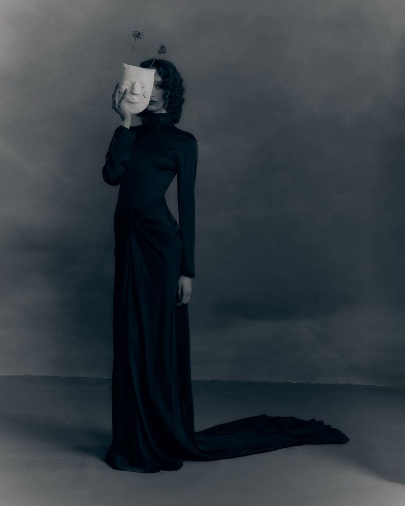 Model in long black dress holding a white mask over her face against a dark studio backdrop for Patrick McDowell AW26 lookbook