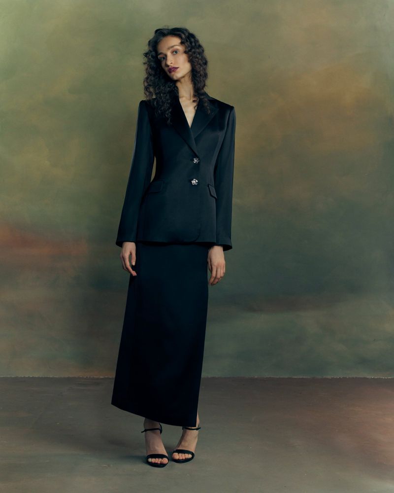 Model in a black suit jacket and matching long skirt standing against an artistic painterly backdrop