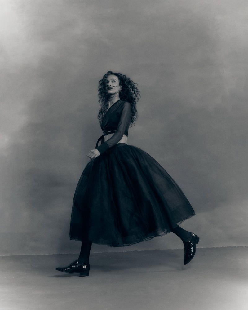 Model with long curly hair wearing a dark midi skirt and top walking across a mottled studio backdrop