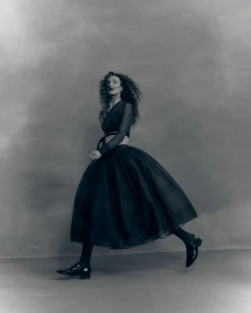 Model with long curly hair wearing a dark midi skirt and top walking across a mottled studio backdrop