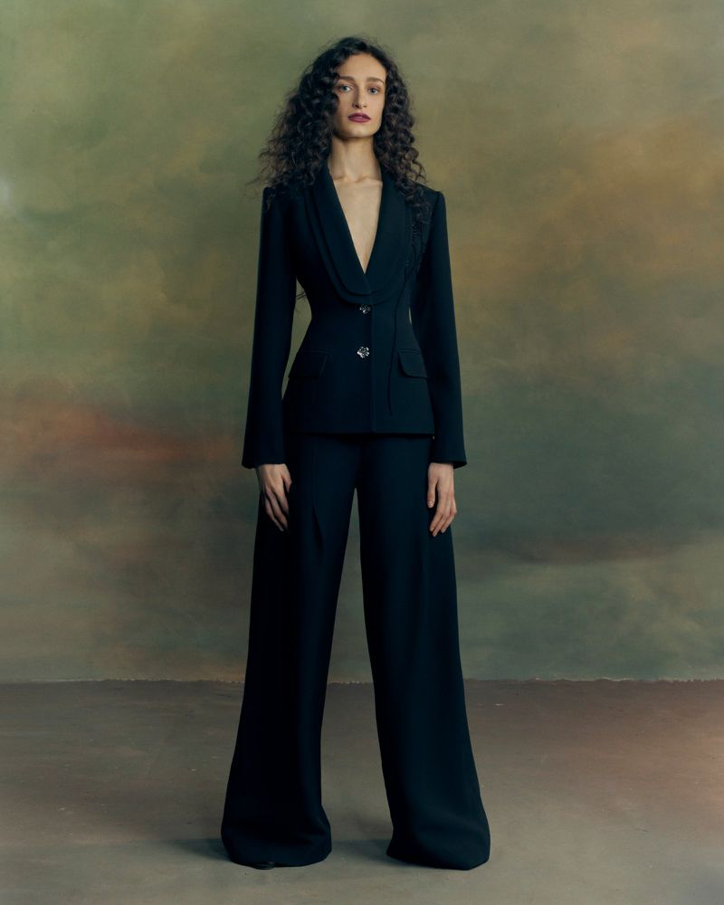 Model with long curly hair wearing a tailored black blazer and matching trousers standing against a painted textured backdrop.