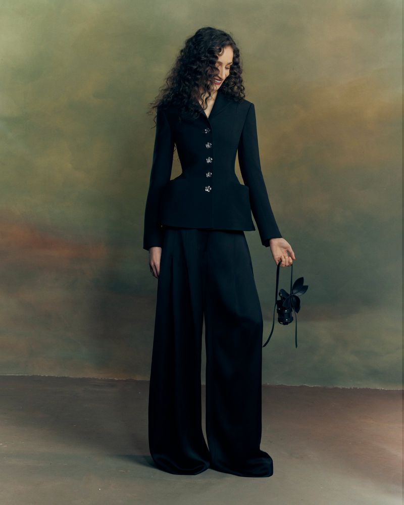 Model with long dark curly hair wearing a black blazer and wide-leg trousers against a textured mottled backdrop.