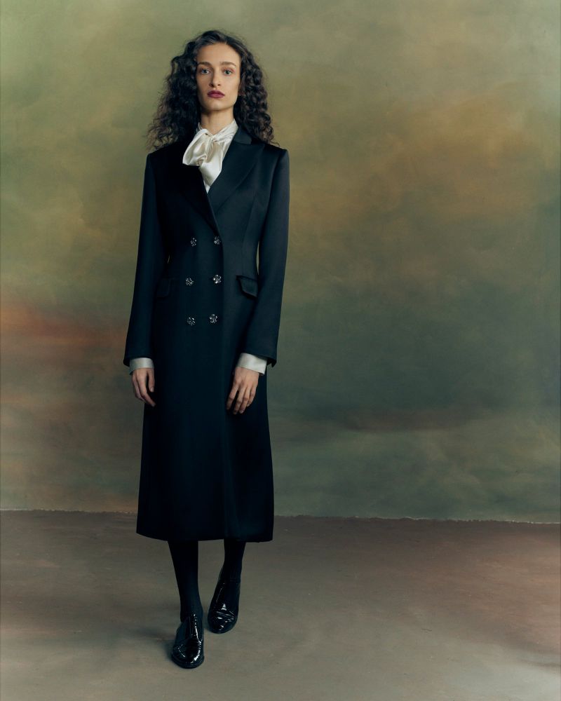 Model with dark curly hair wearing a long black coat and white bow blouse standing against a painted moody backdrop