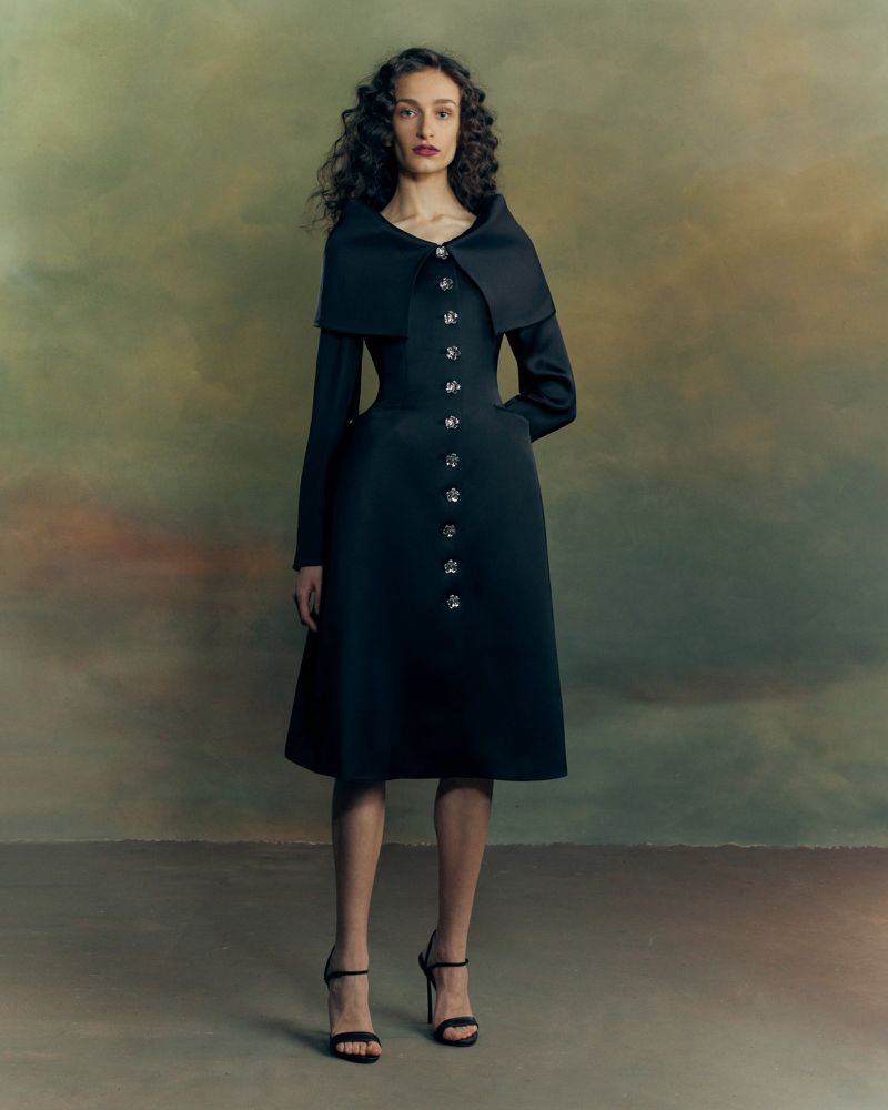 Model wearing a structured black dress with ornate buttons and a wide collar standing against a textured backdrop