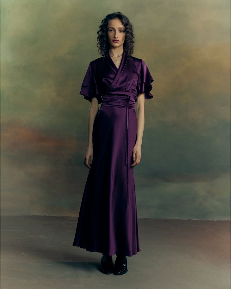 Model with dark curly hair wearing a long purple satin dress with flared sleeves against a painterly textured background.