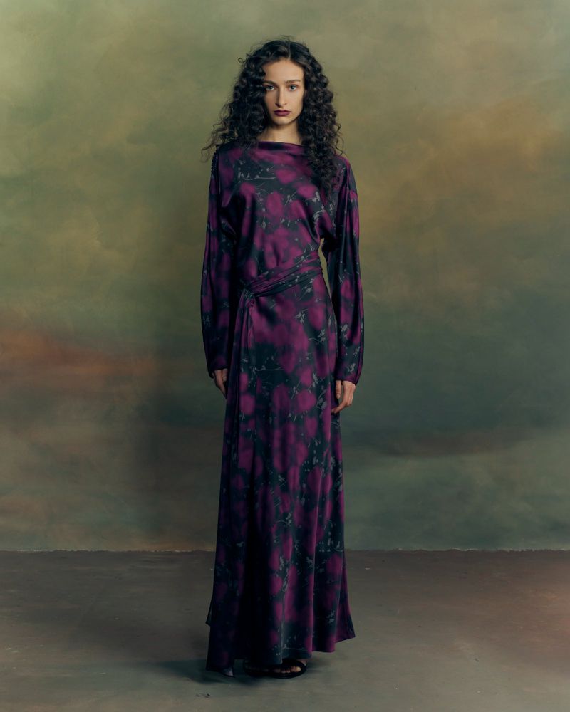 Model with long dark curly hair standing in a purple patterned long-sleeved dress against a moody painted backdrop.