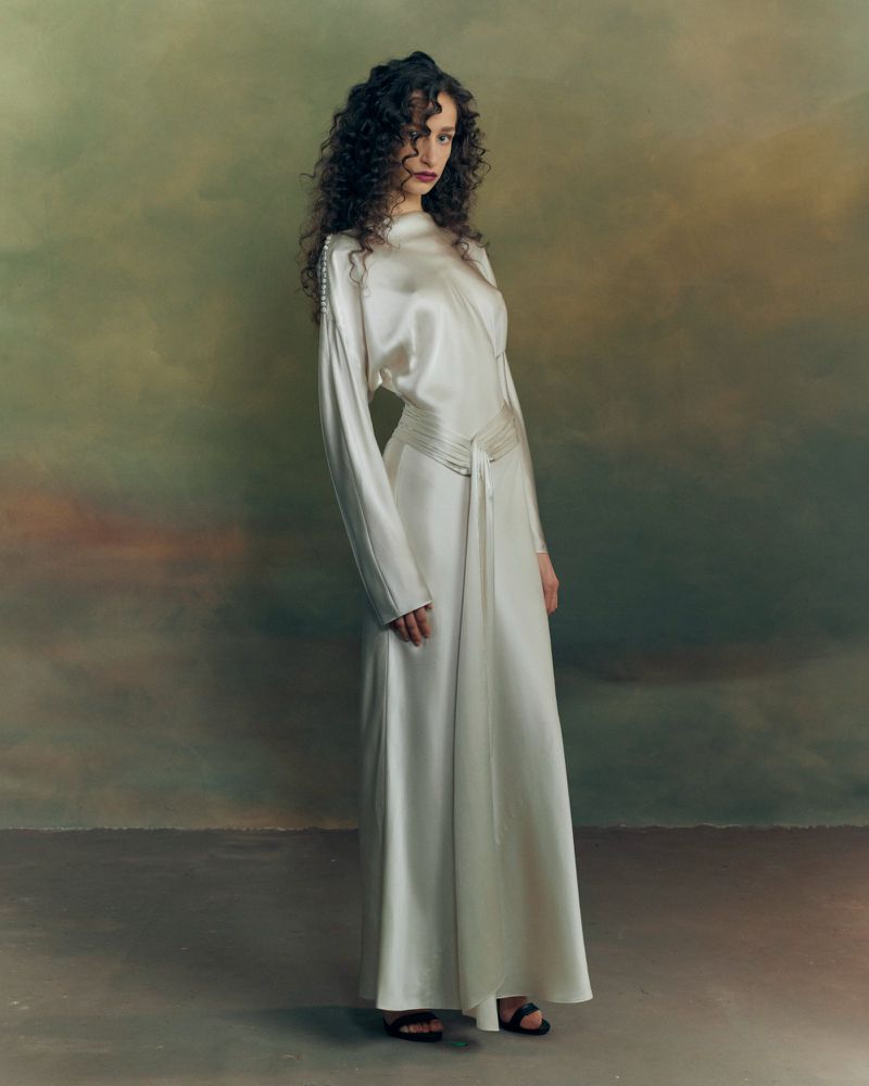 Model with long curly dark hair wearing a long white silk gown standing against a moody painted backdrop