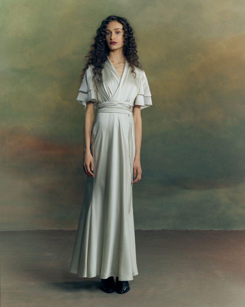 Model with long dark curly hair wearing a pale wrap dress with flared layered sleeves standing against a painterly backdrop.