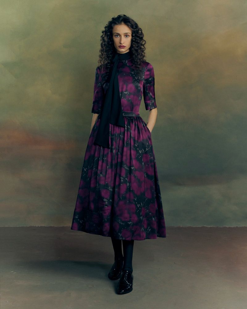 Model with dark curly hair wearing a patterned purple midi dress against a moody painted backdrop.