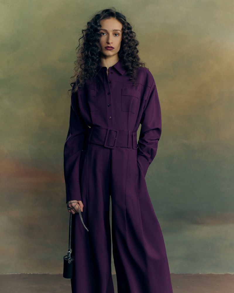 Model with long dark curly hair wearing a deep purple belted jumpsuit holding a small black handbag against a muted painted backdrop