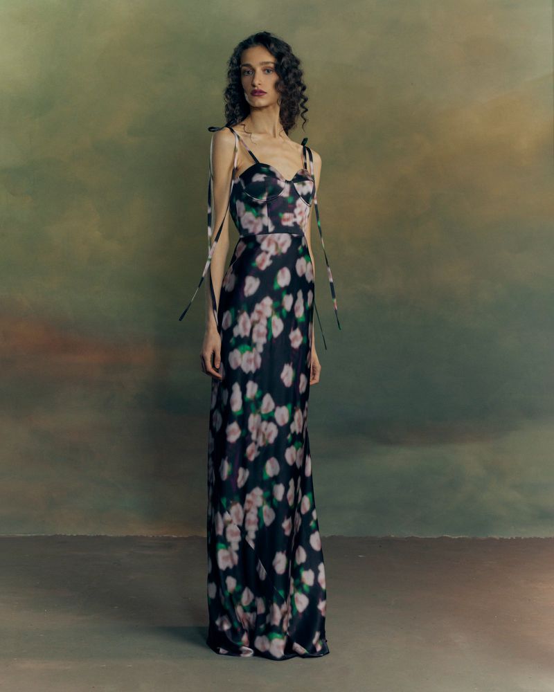 Model with long dark curly hair wearing a dark floral slip dress with shoulder ties against a painterly backdrop.