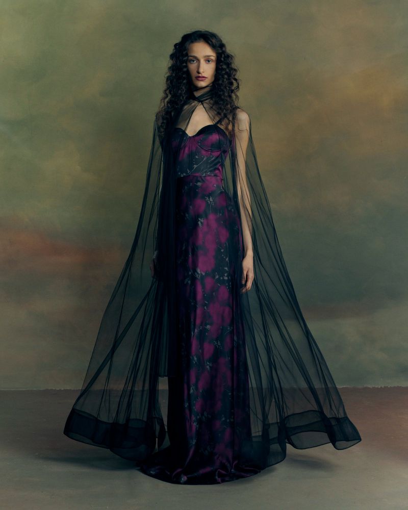 Model wearing a purple patterned gown with a sheer black tulle cape against a painterly textured backdrop.