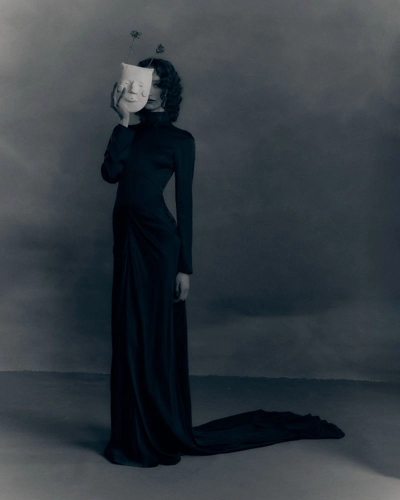 Model in long black dress holding a white mask over her face against a dark studio backdrop for Patrick McDowell AW26 lookbook
