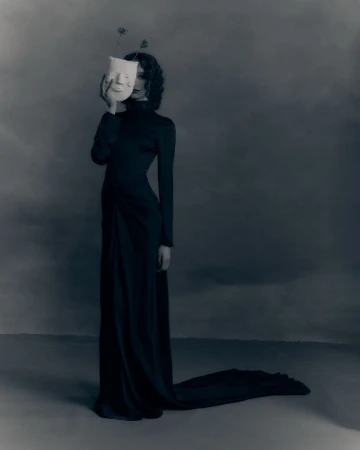 Model in long black dress holding a white mask over her face against a dark studio backdrop for Patrick McDowell AW26 lookbook