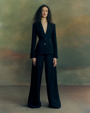 Model with long curly hair wearing a tailored black blazer and matching trousers standing against a painted textured backdrop.