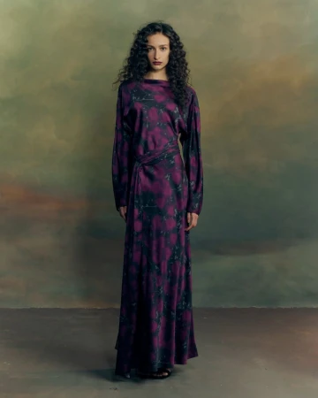 Model with long dark curly hair standing in a purple patterned long-sleeved dress against a moody painted backdrop.