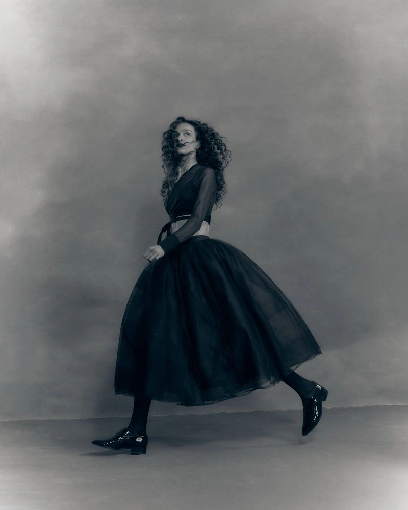 Model with long curly hair wearing a dark midi skirt and top walking across a mottled studio backdrop