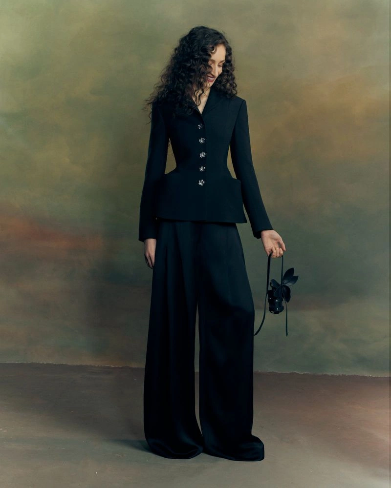Model with long dark curly hair wearing a black blazer and wide-leg trousers against a textured mottled backdrop.
