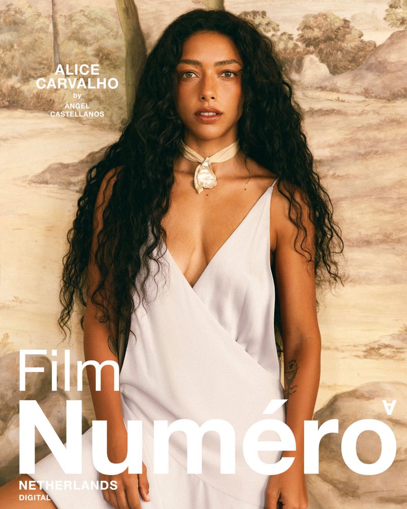 Portrait of Alice Carvalho with long dark curly hair wearing a white slip dress and neck choker against a painted mural backdrop.