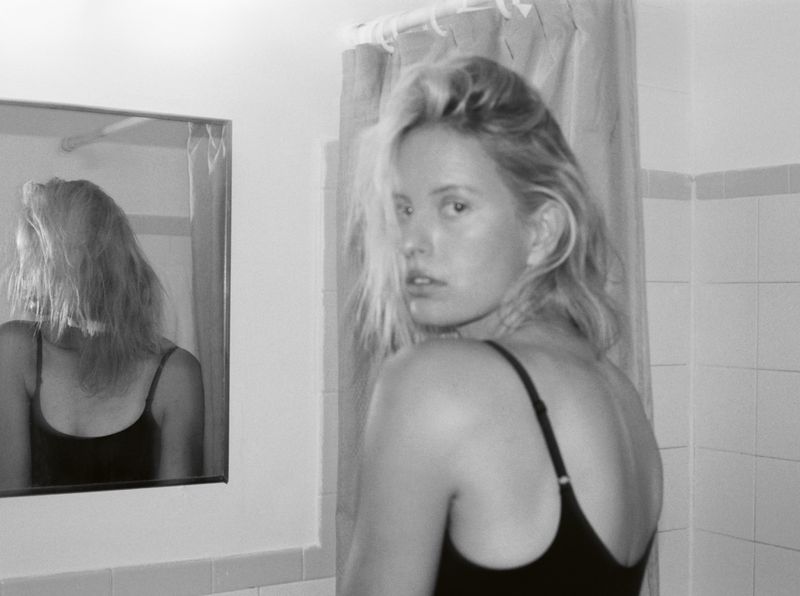 Black and white portrait of a woman in a black tank top looking over her shoulder in a tiled bathroom with a mirror.