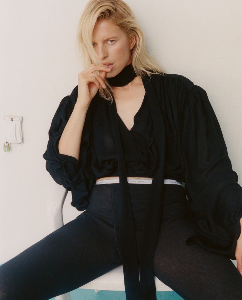 Karolina Kurkova sitting on a chair wearing a black top and pants against a white background