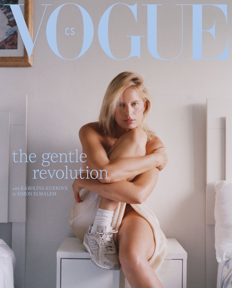 Model Karolina Kurkova seated on a white nightstand wearing white socks and sneakers against a bedroom background
