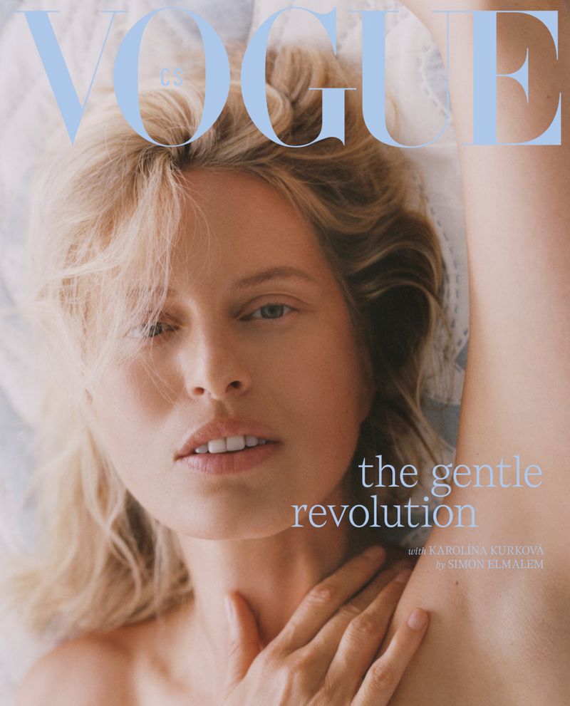 Close-up portrait of model Karolina Kurkova with blonde hair resting her hand on her collarbone against a white textile background.