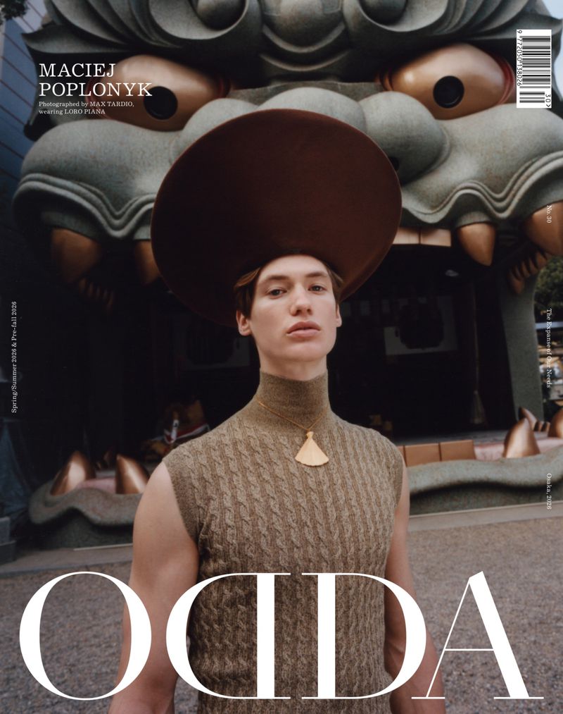 Model wearing a wide-brimmed brown hat and cable-knit sleeveless top against an ornate stone guardian statue background.