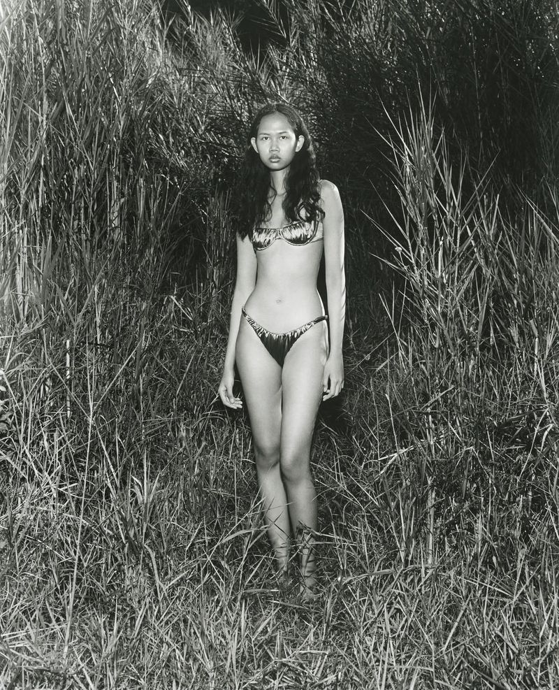 Model standing in tall marsh grass wearing a patterned two-piece swimsuit in a black and white fashion editorial photograph.