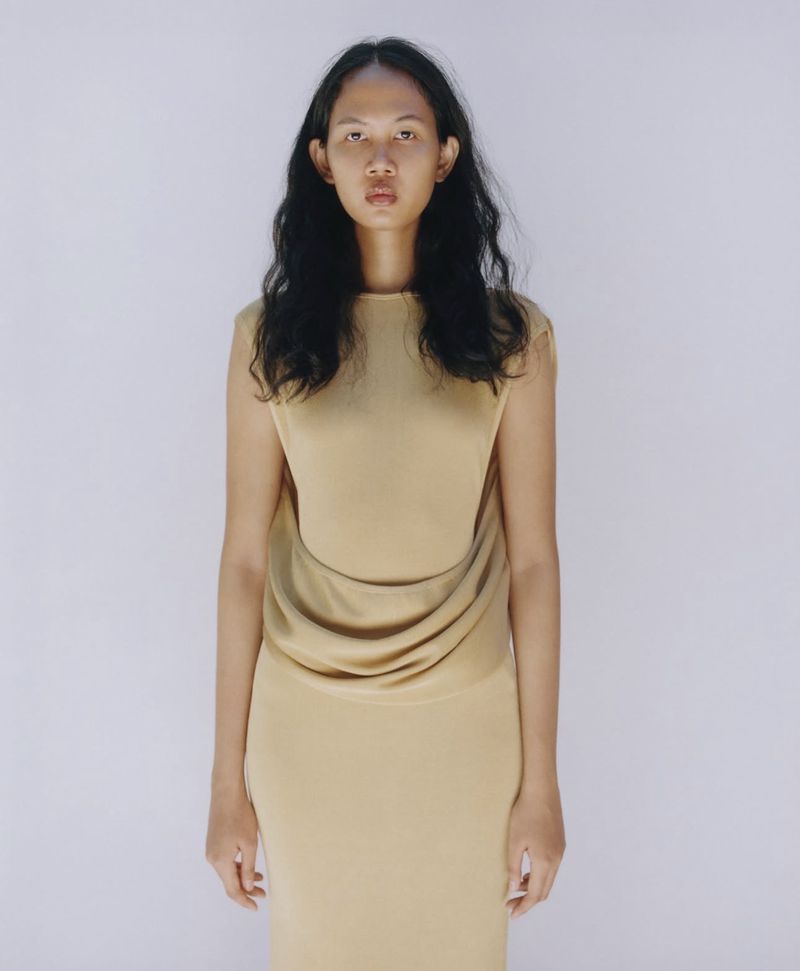 Model with long dark wavy hair wearing a beige sleeveless dress against a plain light background