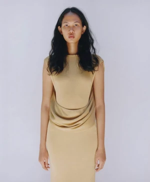 Model with long dark wavy hair wearing a beige sleeveless dress against a plain light background