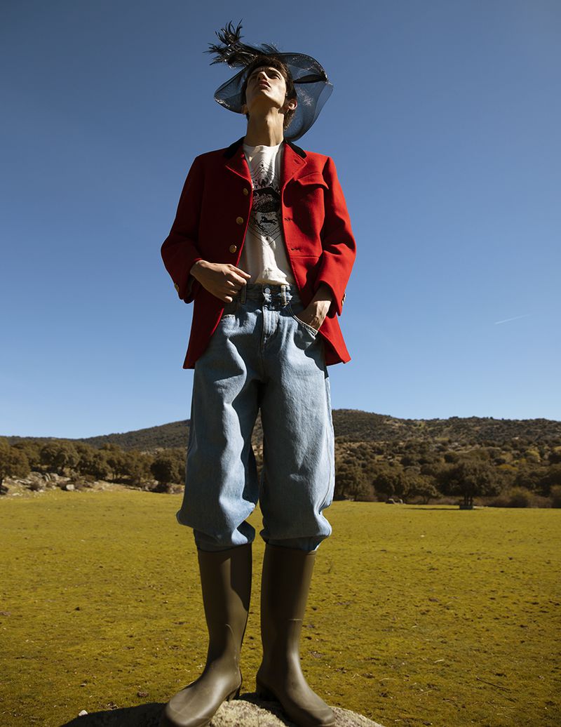 Model standing outdoors wearing a red jacket, light blue jeans, tall boots, and a wide-brimmed hat against a clear blue sky
