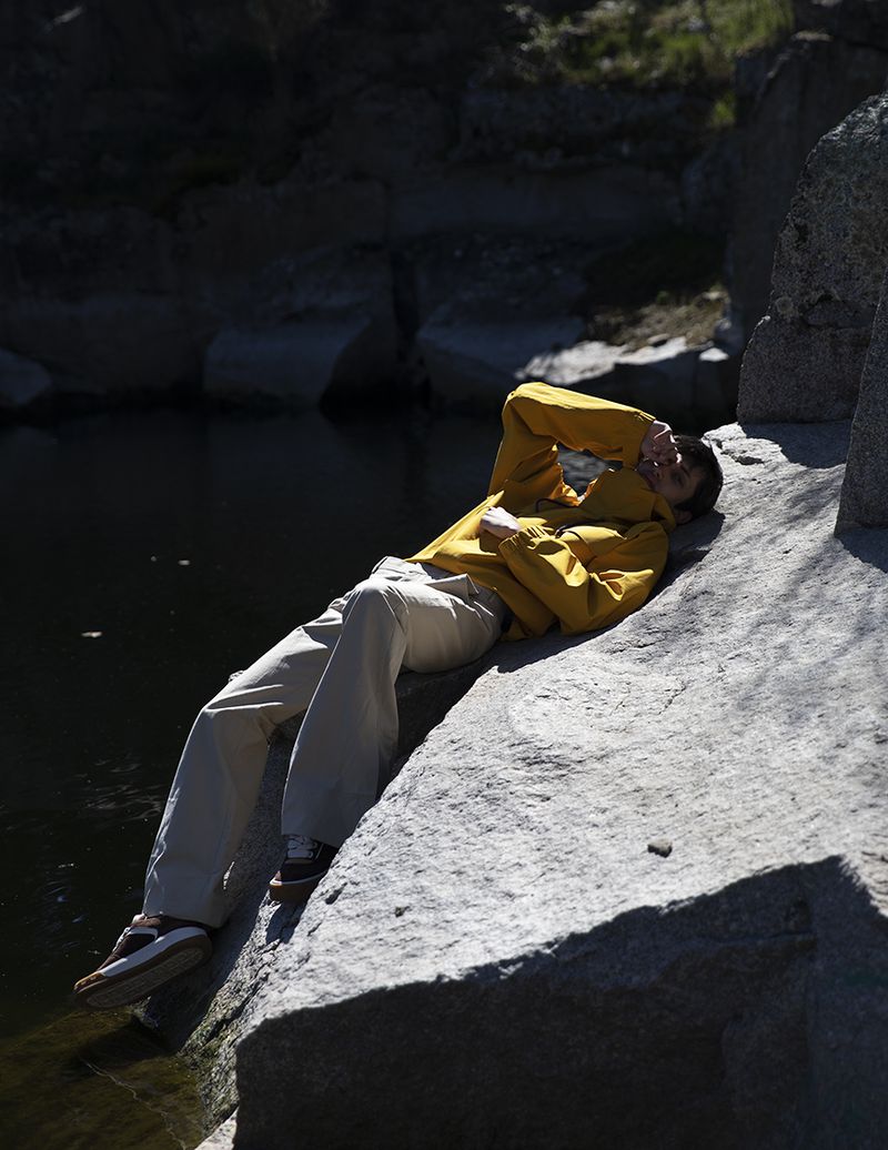 Model wearing a yellow jacket and white trousers lying on a rocky ledge near water for L'Officiel Hommes Malaysia.