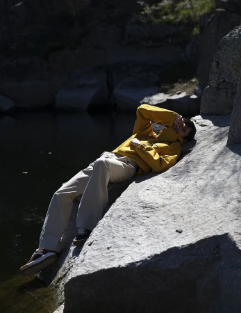 Model wearing a yellow jacket and white trousers lying on a rocky ledge near water for L'Officiel Hommes Malaysia.
