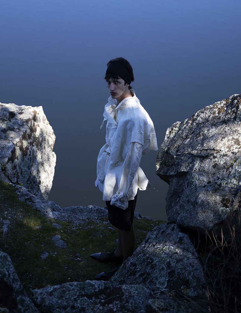 Model wearing a white shirt and dark shorts standing between rocky outcrops overlooking water for L'Officiel Hommes Malaysia.