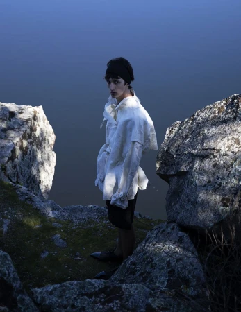 Model wearing a white shirt and dark shorts standing between rocky outcrops overlooking water for L'Officiel Hommes Malaysia.