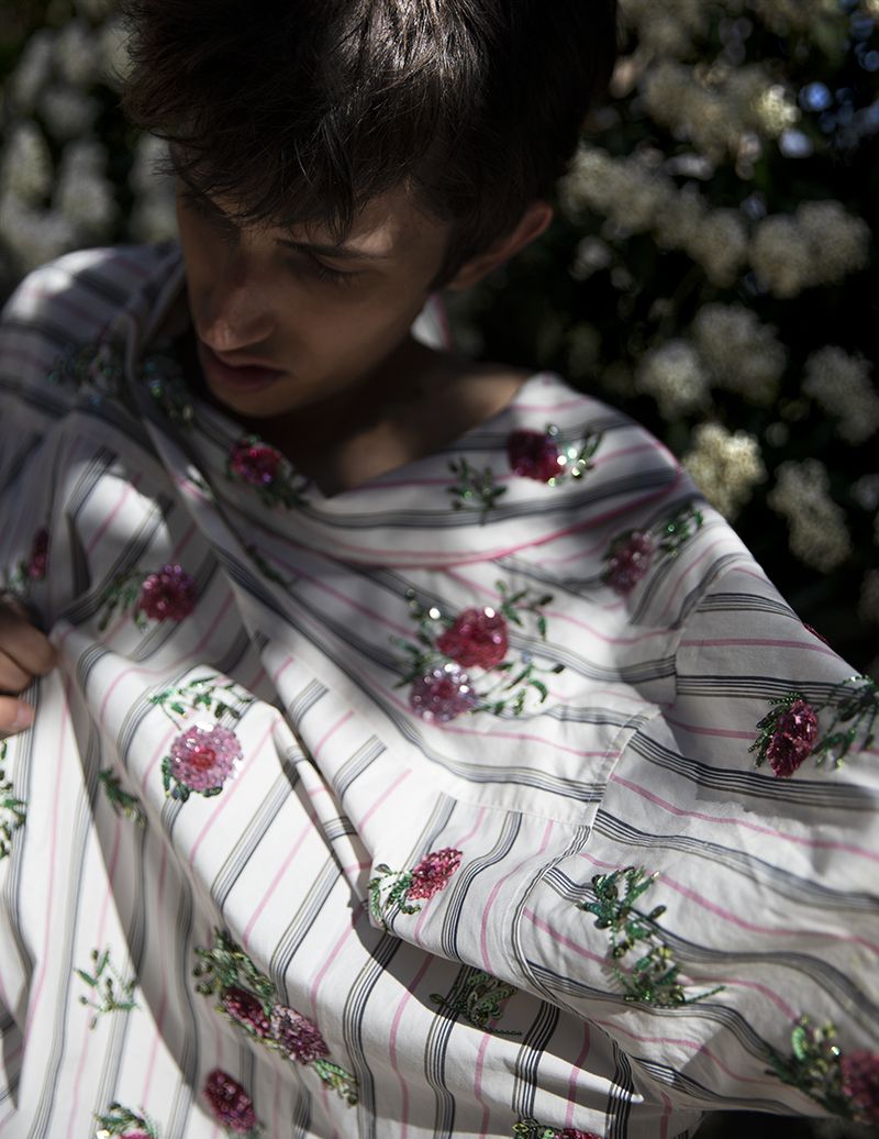 Model wearing a white and pink striped shirt embroidered with pink floral appliqués outdoors among blooming bushes