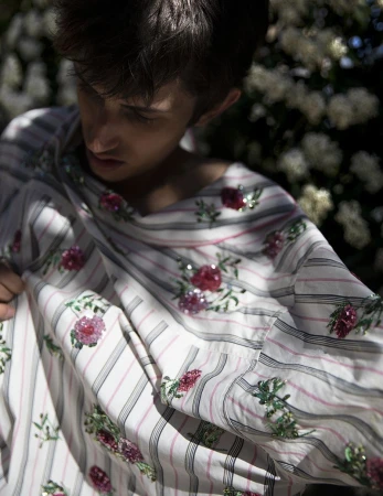 Model wearing a white and pink striped shirt embroidered with pink floral appliqués outdoors among blooming bushes
