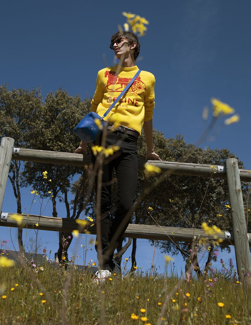 Model wearing a yellow sweater leaning against a wooden fence in a sunny meadow with blue sky