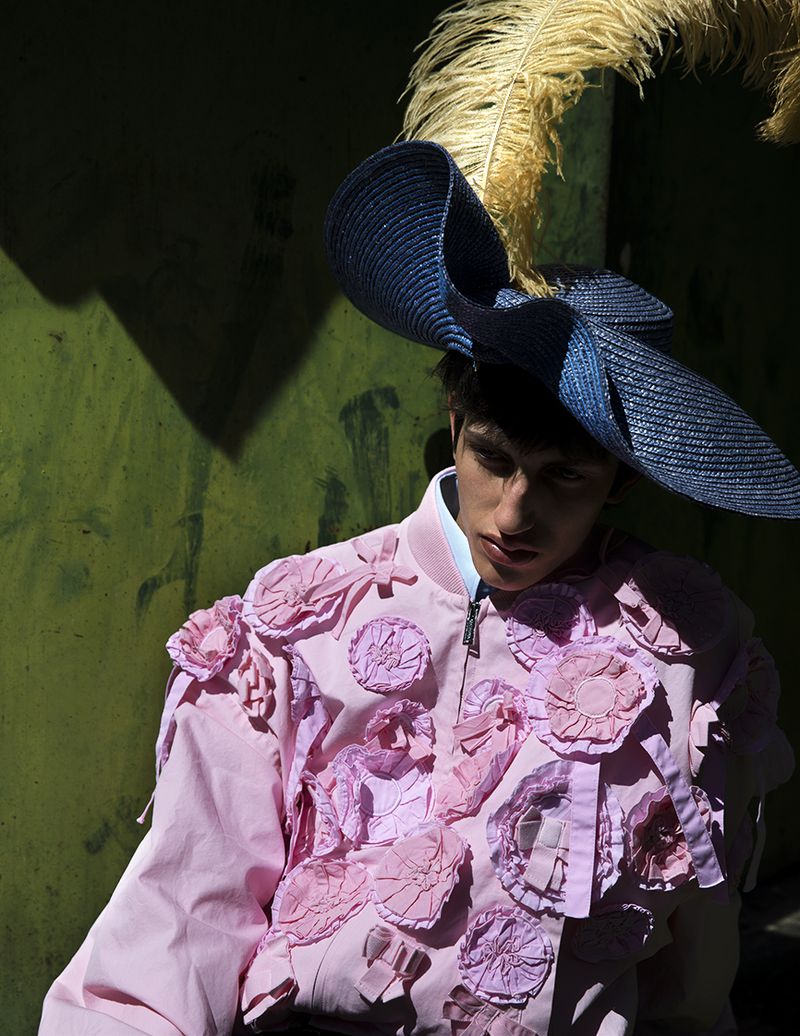 Male model wearing a pink floral jacket and a large blue hat with a feather against a green wall