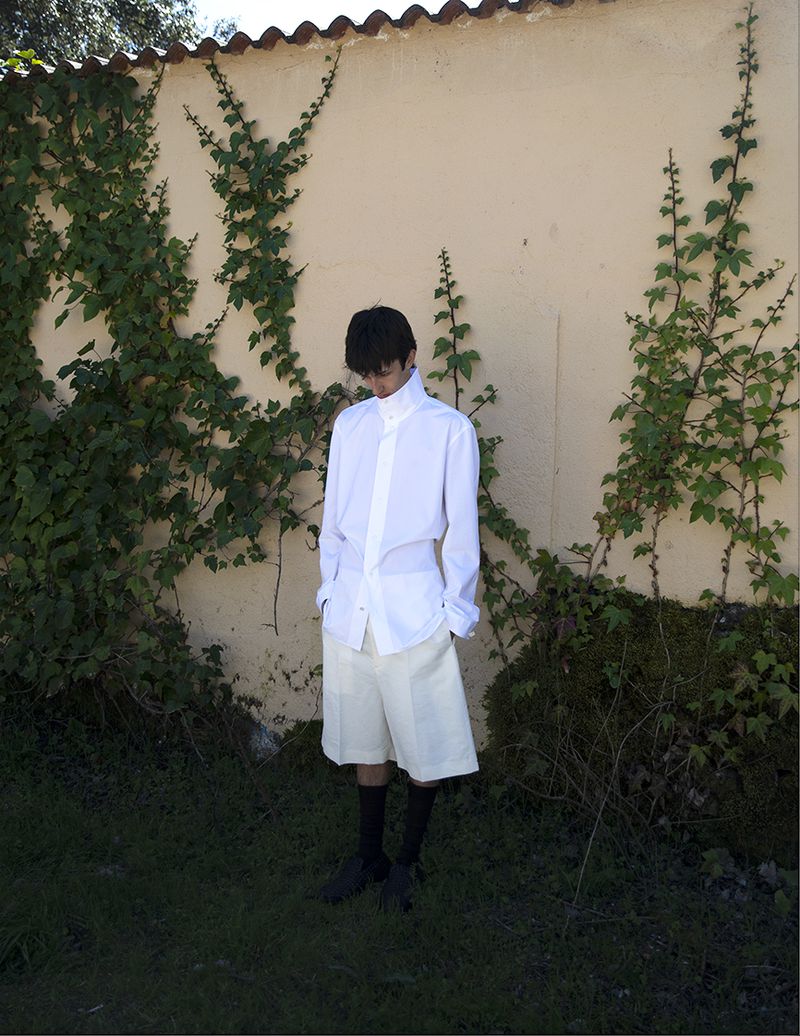Model standing in front of a stucco wall covered in climbing vines, wearing a white collared shirt and light shorts.