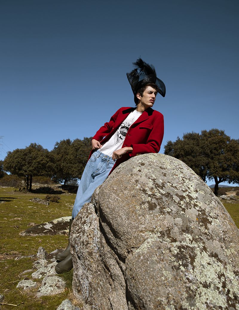 Model wearing a red blazer, graphic t-shirt, light denim jeans, and a black feathered hat leaning against a large rock outdoors.