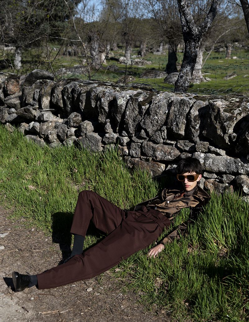 Male model in brown trousers and silk shirt lying on grass against a stone wall