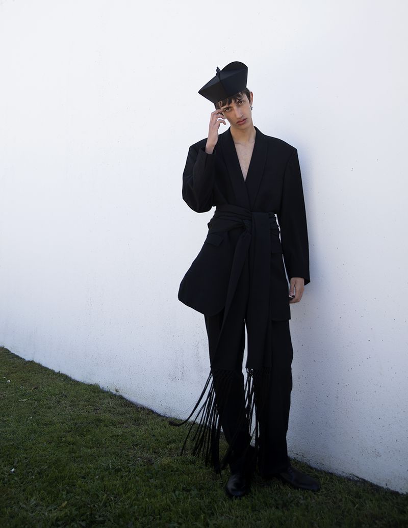 Model Nicolas Secades in a black suit with a brimless hat posing against a white wall for L'Officiel Hommes Malaysia.